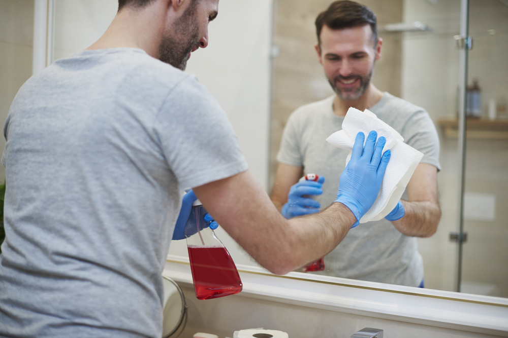 Persona limpiando el espejo de un baño con guantes de goma y limpiador en spray, asegurando un acabado brillante y sin manchas.